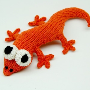 May include: A hand-knitted orange lizard toy with white eyes. The lizard has a long tail and four legs.