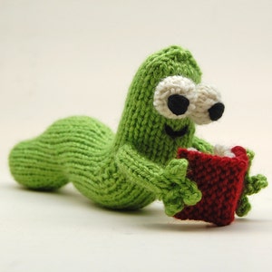 May include: A green knitted worm toy holding a red knitted book. The worm has white eyes and a smiling mouth.