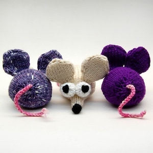 May include: Three knitted mice toys. One is purple, one is beige with black eyes, and one is a purple and white speckled pattern. All have pink tails.
