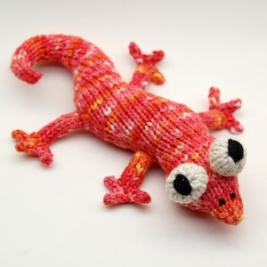 May include: A knitted gecko toy with a red, orange and white colour scheme. The gecko has large, white eyes with black pupils and a curled tail.