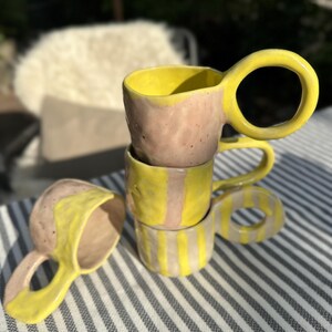 Könnte beinhalten: Ein Stapel von vier handgefertigten Keramikbechern. Die Becher haben ein gelbes Innenleben und einen Griff, die mit einem rosafarbenen Außenbereich kontrastieren. Ein Becher liegt auf der Seite. Die Becher stehen auf einer gestreiften Oberfläche.