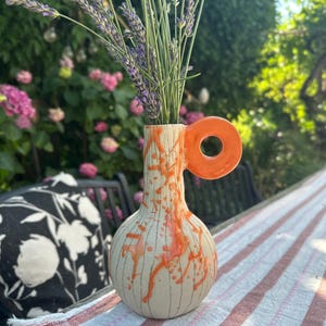 Könnte beinhalten: Eine Keramikvase mit rundem Boden und engem Hals, die einen leuchtend orangefarbenen, kreisförmigen Henkel aufweist. Die cremeweiße Oberfläche ist mit dünnen vertikalen schwarzen Linien und unregelmäßigen orangefarbenen Farbspritzern verziert.