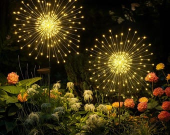 May include: Two solar-powered, starburst-shaped garden lights illuminate a nighttime garden scene. The lights emit a warm yellow glow, resembling fireworks. The garden features green foliage and orange and yellow flowers.