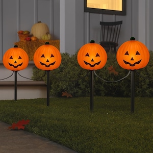 May include: A set of four orange jack-o'-lantern pumpkin pathway lights on black stakes. Each pumpkin features a carved face with triangular eyes, a triangular nose, and a wide, jagged mouth. The lights are connected by a black electrical cord.