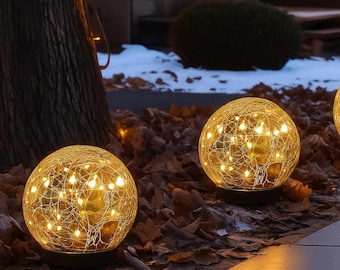 May include: Three illuminated spherical decorative lights. Each sphere is encased in a web-like design, with warm white lights visible within. The lights are set on a dark base, resting on a bed of fallen leaves. The background includes a tree trunk and a snowy area.