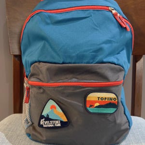 May include: A blue and gray backpack with red accents and two patches. The patches are for "Tofino" and "Revelstoke National Park".