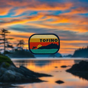 Può includere: Una toppa in stile retrò con una scena di tramonto, un delfino che salta sopra un'onda e la parola "TOFINO" su uno stendardo giallo. La toppa è su uno sfondo sfocato di una spiaggia al tramonto.