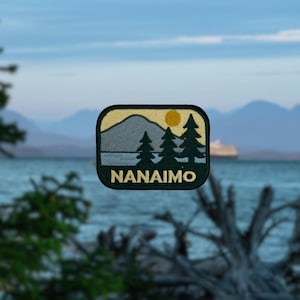May include: A rectangular patch featuring a landscape scene with the word "NANAIMO". The patch has a yellow border and a design of a mountain, trees, and a sun. The background shows a body of water and mountains.