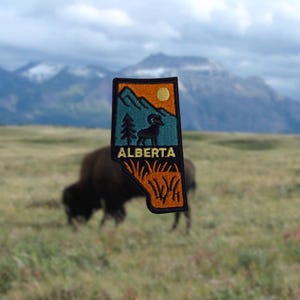 Könnte beinhalten: Gestickter Aufnäher in Form von Alberta, Kanada, mit einer Berglandschaft, einem Widder, Kiefern und einer Sonne. Das Wort "ALBERTA" ist weiß gestickt. Der Aufnäher ist schwarz und orange umrandet.