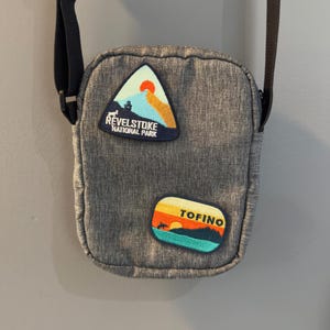 May include: A gray crossbody bag with two embroidered patches. The top patch is a triangle with the text "Revelstoke National Park". The bottom patch is a rectangle with the text "Tofino".