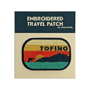 May include: Embroidered travel patch with a sunset scene and a dolphin jumping out of the water. The patch says "TOFINO" in black text.