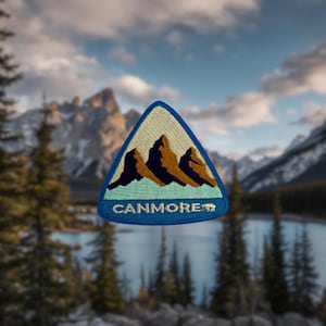 May include: A blue triangular patch with a white background and an embroidered image of three mountains. The text "CANMORE" is embroidered in blue below the mountains. The patch is in front of a blurred background of mountains and a lake.