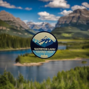 May include: A blue and white embroidered patch featuring the text "Waterton Lakes National Park" and a mountain range design. The patch is in focus against a blurred background of a lake and mountains.