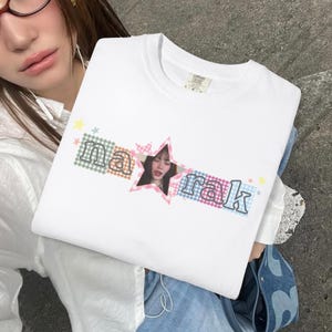 Puede incluir: Una camiseta blanca de cuello redondo con un diseño gráfico que incluye el texto "na rak" en una fuente de contorno oscuro. Las letras se sitúan sobre cuadros vichy de colores pastel, con una estrella rosa que contiene un retrato fotográfico.
