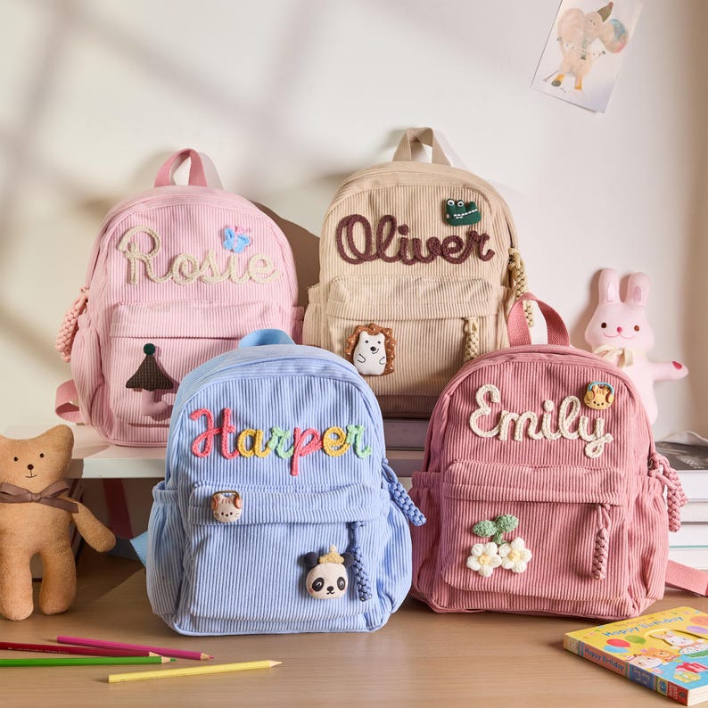K&ouml;nnte beinhalten: Vier Cordrucks&auml;cke in Rosa, Beige und Blau, jeweils mit einem aufgestickten Namen auf der Vorderseite: Rosie, Oliver, Harper und Emily. Die Rucks&auml;cke haben dekorative Aufn&auml;her und eine Vordertasche. Ideal f&uuml;r Kinder.