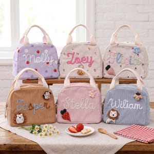 May include: Six corduroy lunch bags in pastel shades, each embroidered with a name. The bags have a top handle, zip closure, and decorative elements. The bags are displayed on a wooden surface, with a plate of strawberries.