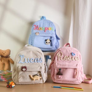 K&ouml;nnte beinhalten: Drei Cord-Rucks&auml;cke in Blau, Beige und Rosa, jeweils mit einem gestickten Namen personalisiert. Der blaue Rucksack hat einen Panda-Aufn&auml;her, der beige einen B&auml;ren und Dinosaurier, und der rosa einen Pilz und Schmetterling.