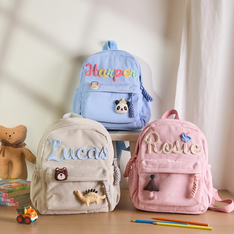 K&ouml;nnte beinhalten: Drei Cord-Rucks&auml;cke in Blau, Beige und Rosa, jeweils mit einem gestickten Namen personalisiert. Der blaue Rucksack hat einen Panda-Aufn&auml;her, der beige einen B&auml;ren und Dinosaurier, und der rosa einen Pilz und Schmetterling.