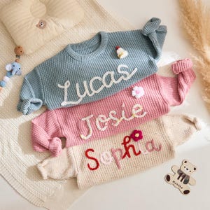 May include: Three personalized baby sweaters in blue, pink, and cream. Each sweater has a name embroidered in a contrasting color: "Lucas", "Josie", and "Sophia". Decorative floral and holiday accents are also present.