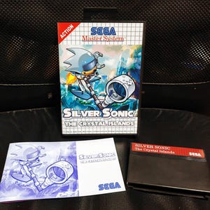 May include: A Sega Master System game cartridge for Silver Sonic: The Crystal Islands. The cartridge is black with a red label that reads "Silver Sonic The Crystal Islands" and the Sega logo. The front cover of the game is blue and features a silver robot character with a drill.