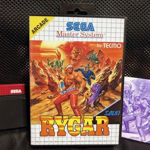 Puede incluir: Cartucho de juego SEGA Master System para Rygar. La carátula presenta un guerrero musculoso con un escudo, luchando contra criaturas fantásticas. El título del juego está en letras grandes y amarillas. El cartucho es negro y rojo.
