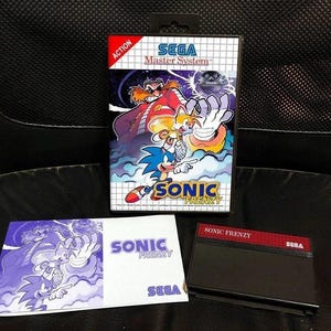 May include: A Sega Master System game cartridge for Sonic Frenzy. The cartridge is black with red accents and the game title is printed in white. The game box art features Sonic the Hedgehog, Tails, and Dr. Robotnik. The box art is blue, yellow, and red.