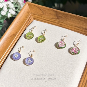 Op de afbeelding: Zes paar handgemaakte oorbellen met goudkleurige haken. De oorbellen hebben heldere, ronde harsbollen met gedroogde bloemen in groen, paars en roze. De tekst "MemesoHouse Handmade Jewelry" is zichtbaar.