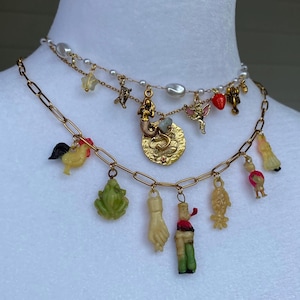 May include: A two-layered necklace with gold chains and a variety of charms. The top layer has pearls and small charms, while the bottom layer features a rooster, frog, hand, and other whimsical figures. The charms are in various colours.