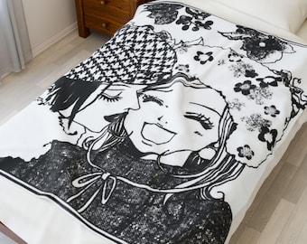 May include: A white blanket featuring a black and white manga-style illustration of two women. One woman is kissing the other, who is smiling. The blanket has a black and white houndstooth pattern and floral accents.