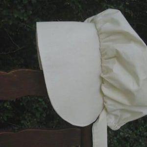 May include: A cream-colored bonnet with a wide brim and gathered fabric at the back. The bonnet is made of a soft, light-colored material and is tied with a matching ribbon. The bonnet is resting on a wooden chair.