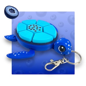 May include: A blue turtle keyring with a light blue shell and a silver clasp. The turtle has white polka dots on its head and a round eye. A blueberry is floating in the upper left corner.