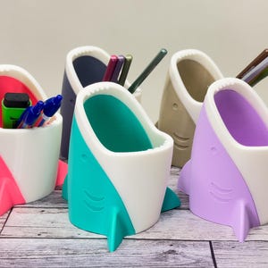 Puede incluir: Cinco organizadores de escritorio con forma de tiburón de diferentes colores hechos de plástico. Cada organizador tiene una barriga blanca y una parte superior de color diferente: rosa, verde azulado, gris, beige y morado. Los organizadores están llenos de bolígrafos y lápices.