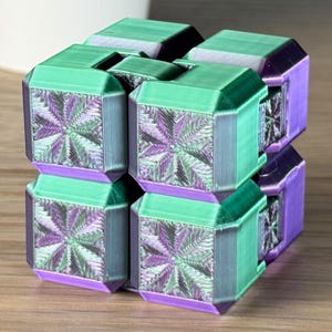 Puede incluir: Un cubo de rompecabezas impreso en 3D con un esquema de color verde y morado. El cubo está formado por cubos más pequeños que están conectados por bisagras. Cada cubo tiene un patrón en forma de estrella en su superficie.