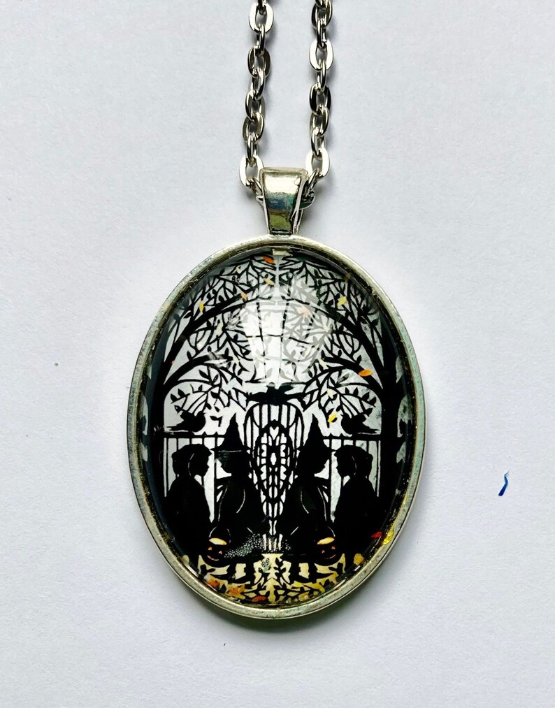 May include: A silver chain necklace with a round glass pendant featuring a black and white silhouette of three figures standing in front of a gate with a tree behind them. The figures are wearing hats and the gate is decorated with a design.