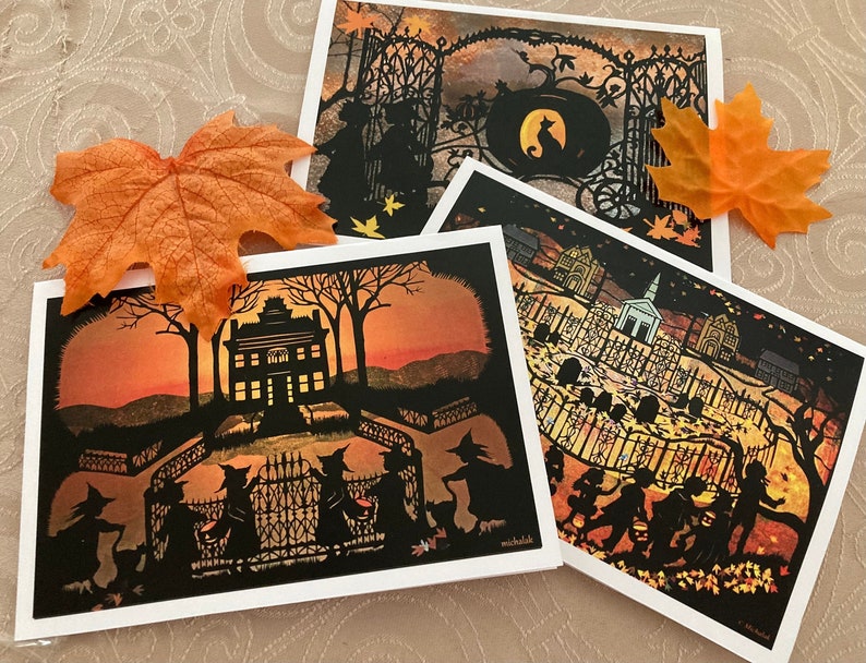 May include: Three Halloween-themed cards with intricate paper cutouts. The first card features a silhouette of a house with three witches in front of it. The second card shows a silhouette of a graveyard with a church in the background. The third card depicts a silhouette of a cat sitting in front of a gate.