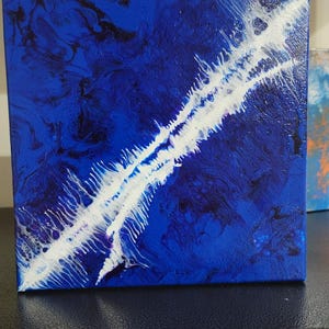 Könnte beinhalten: Quadratische Leinwandkunst mit einem lebendigen abstrakten Design. Das Kunstwerk zeigt einen tiefblauen Hintergrund mit wirbelnden Mustern in Schwarz und Weiß. Ein kräftiger weißer Streifen schneidet durch die Mitte und erzeugt einen dynamischen visuellen Effekt. Das Stück wird auf einer dunklen Oberfläche präsentiert.
