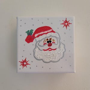 Pode incluir: Uma pintura em tela branca do Papai Noel com um chapéu vermelho e barba branca. O fundo é branco com glitter prateado e flocos de neve vermelhos.