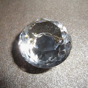 May include: A clear, faceted crystal paperweight with a rounded shape. The crystal reflects light, creating a sparkling effect. The paperweight is sitting on a textured, gray surface.