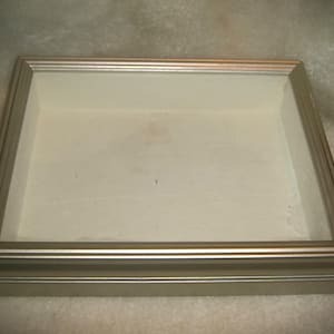 May include: A rectangular display case with a silver-colored frame and a clear glass top. The interior is a light cream color. The case is sitting on a light-colored surface.