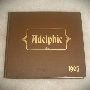 May include: A brown book with the word "Adelphic" in gold lettering inside a gold rectangular frame. The year "1907" is printed in gold below the frame. The book has a slightly worn appearance.