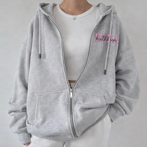 May include: A light gray zip-up hoodie with a hood and front pockets. The word "Kathleen" is embroidered in pink on the upper left chest. The hoodie is worn over a white top and paired with white pants.