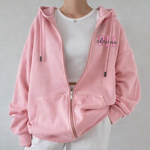 May include: A light pink zip-up hoodie with a hood and front pockets. The hoodie has the text "011 abrams" printed on the upper right chest area. The model is wearing a white top and white pants.