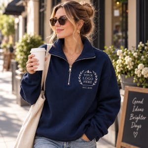 May include: A navy blue quarter-zip sweatshirt featuring a white embroidered laurel wreath design on the left chest. The embroidery includes the text "YOUR LOGO HERE" and "EST. 2026" with a small mountain graphic centered above the text.