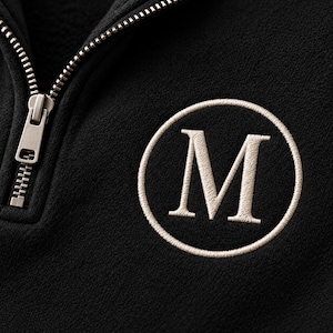 May include: A black fleece quarter-zip pullover featuring a metallic silver zipper and a circular embroidered patch on the chest. The patch displays a large, cream-colored capital letter M centered within a stitched circular border.