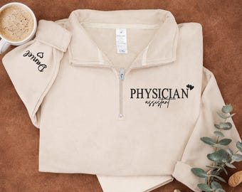 May include: A beige quarter-zip sweatshirt with the text "PHYSICIAN ASSISTANT" in black. The sleeve cuff has the name "Daniel" and a heart symbol. A white mug with coffee is on the left.