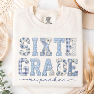 May include: A cream-colored crewneck sweatshirt featuring the text SIXTH GRADE in large, block-style letters filled with various blue and white patterns, including gingham, floral, and polka dot prints, with the name mi parker written below.