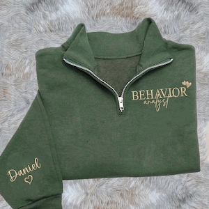 May include: A dark green quarter-zip sweatshirt with the words "BEHAVIOR analyst" embroidered in a gold script font. The sleeve has the name "Daniel" embroidered in gold script with a heart symbol. The sweatshirt has a silver zipper.