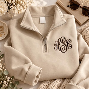 May include: A beige quarter-zip pullover sweatshirt featuring a dark brown embroidered monogram with the letters ASC in a decorative script font on the left chest. The garment has a high collar, a silver-toned metal zipper, and long sleeves.
