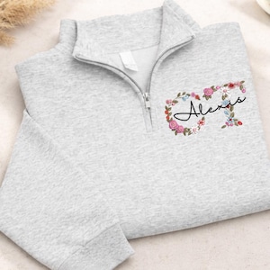 May include: A light gray quarter-zip sweatshirt with the name "Alexis" in cursive script, surrounded by a floral wreath of pink, red, and blue flowers. The sweatshirt has a ribbed cuff and a white tag.