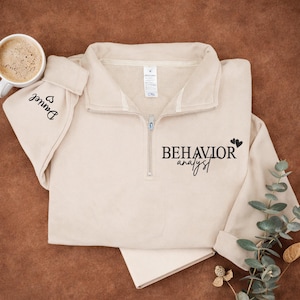 May include: A beige quarter-zip sweatshirt with the words "BEHAVIOR analyst" printed in black. The sleeve cuff has the name "Daniel" printed in black with a heart symbol. A mug of coffee sits next to the sweatshirt.